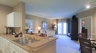 This single-story residence is designed with an open-concept floor plan that promotes a natural flow between the kitchen, dining, and living spaces. The kitchen features a wraparound counter with a double-basin sink, light-toned countertops, and a breakfast bar that opens directly to the main living area, making it ideal for entertaining or casual dining.

Crown molding runs along the ceilings, adding a refined architectural detail throughout the space. The living area is finished with plush neutral carpeting and benefits from abundant natural light streaming through a set of French doors, which lead to an outdoor area and enhance the sense of openness. The French doors are framed by decorative curtains that add a layer of visual warmth.

A hallway visible from the main area leads to a bedroom, which appears bright and comfortably sized. The overall color palette consists of warm beige and taupe tones, contributing to a cohesive and inviting ambiance. The layout maximizes functionality while maintaining an airy, well-connected feel across all areas of the space.