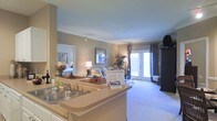 This single-story residence is designed with an open-concept floor plan that promotes a natural flow between the kitchen, dining, and living spaces. The kitchen features a wraparound counter with a double-basin sink, light-toned countertops, and a breakfast bar that opens directly to the main living area, making it ideal for entertaining or casual dining.

Crown molding runs along the ceilings, adding a refined architectural detail throughout the space. The living area is finished with plush neutral carpeting and benefits from abundant natural light streaming through a set of French doors, which lead to an outdoor area and enhance the sense of openness. The French doors are framed by decorative curtains that add a layer of visual warmth.

A hallway visible from the main area leads to a bedroom, which appears bright and comfortably sized. The overall color palette consists of warm beige and taupe tones, contributing to a cohesive and inviting ambiance. The layout maximizes functionality while maintaining an airy, well-connected feel across all areas of the space.