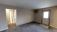 This single-floor, two-bedroom, one-bathroom residence offers a functional and straightforward layout designed for comfortable everyday living. The living area features neutral-toned walls and grey carpeting that extends throughout the main spaces, creating a cohesive and understated aesthetic. A baseboard heating system runs along the perimeter of the living room, ensuring efficient warmth during colder months, while a wall-mounted air conditioning unit provides climate control year-round. Natural light enters through a windowed entry door, brightening the space and giving the front entryway a welcoming feel. The entry includes a tiled threshold, providing a practical transition zone from the outdoors. A hallway visible from the living area leads to the bedrooms and bathroom, offering a clear and organized flow through the unit. The neutral color palette of beige and grey tones throughout lends itself well to a variety of furnishing styles, making it easy to personalize. Overall, this residence provides a clean, move-in-ready living environment with essential amenities, practical climate control, and a simple, efficient floor plan suited for individuals or small households.