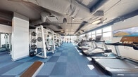 This modern fitness center spans a single floor and offers a comprehensive, thoughtfully organized workout environment. The space is divided into two distinct zones, seamlessly connected by full-height glass partition walls and a central glass door, creating an open and airy feel while allowing visual continuity throughout.

The main workout area features a robust strength training section equipped with weight racks, barbells, plate-loaded machines, and adjustable benches, complemented by a full dumbbell rack displayed in front of floor-to-ceiling mirrors. A dedicated cardio zone lines the perimeter, housing a row of treadmills, elliptical machines, stair climbers, and stationary cycling bikes — each positioned to face large, floor-to-ceiling windows that invite plentiful natural light and outdoor views.

The second room serves as a dedicated studio space, outfitted with stability balls and additional cardio equipment, making it well-suited for group fitness or focused training sessions. Underfoot, a durable blue and grey checkerboard-patterned carpet tile flooring adds both practicality and visual character throughout. Overhead, an exposed industrial ceiling with visible ductwork, pendant lighting, and track fixtures lends the space a contemporary, urban aesthetic. The abundance of natural light streaming through the expansive windows creates a bright and energizing atmosphere ideal for an active workout environment.