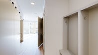 This thoughtfully laid-out one-bedroom, one-bathroom unit offers a clean, modern aesthetic throughout its single floor. Light wood-look flooring flows consistently through every room, creating a cohesive and warm interior. The entryway features a built-in nook with a coat hook and bench, providing a practical landing space upon entry. A hallway connects the living areas to the private spaces, with dark-paneled interior doors adding a subtle contrast to the bright white walls and trim. The bedroom is bathed in natural light thanks to a large floor-to-ceiling sliding glass door that opens to a private balcony, offering views of the surrounding building exterior. The room's clean lines and neutral palette make it a versatile and serene retreat. A dedicated walk-in closet serves the bedroom, outfitted with white wire shelving systems including hanging rods and a multi-tiered shelf unit, maximizing storage efficiency. The bathroom is finished with wood-grain cabinetry topped with a dark countertop, a full glass-door shower enclosure, and stainless steel grab bars, reflecting both style and accessibility considerations. A small utility access panel is also present within the unit. Overall, the space presents a bright, functional, and modern living environment.
