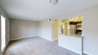 This single-floor residence offers a clean, open-concept design that seamlessly connects the main living area to the kitchen. The spacious living room features soft, neutral-toned carpeting and smooth white ceilings with flush-mount lighting, creating a bright and welcoming atmosphere. Neutral beige walls throughout provide a versatile backdrop suitable for a variety of décor styles. A pass-through counter with a granite surface elegantly separates the living space from the kitchen, maintaining an open and airy feel while defining each functional zone. The kitchen is well-appointed with dark cabinetry, stainless steel appliances including a full-size refrigerator, and track lighting overhead that illuminates the space effectively. Hardwood-style flooring in the kitchen contrasts nicely with the carpeted living area, adding visual interest and practicality. A hallway visible beyond the living room suggests additional rooms within the unit. The overall ambiance is clean, modern, and move-in ready, with a straightforward layout that maximizes usability and comfort throughout the single-level floor plan.