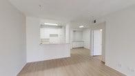 This single-floor space offers a clean, minimalist aesthetic defined by its all-white walls and ceilings paired with light wood-style laminate flooring that runs throughout. The open-concept layout connects the main living area to the kitchen via a raised pass-through counter, which serves as both a functional workspace and a subtle visual divider without fully closing off the space. The kitchen features white cabinetry along the upper and lower sections, a sink positioned at the pass-through, and overhead fluorescent lighting that keeps the area well-illuminated. Ceiling-mounted vents are visible throughout, indicating a central HVAC system. Beyond the main living area, the space continues into a secondary zone with additional low-profile white cabinetry or built-in storage, providing practical utility. A white paneled door leads to a separate room, adding privacy and versatility to the floor plan. The overall ambiance is bright, airy, and neutral, making it a highly adaptable space suited to a variety of uses. The simple, uncluttered design and consistent color palette create a cohesive and functional environment throughout.