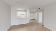 This single-floor space offers a clean, minimalist aesthetic defined by its all-white walls and ceilings paired with light wood-style laminate flooring that runs throughout. The open-concept layout connects the main living area to the kitchen via a raised pass-through counter, which serves as both a functional workspace and a subtle visual divider without fully closing off the space. The kitchen features white cabinetry along the upper and lower sections, a sink positioned at the pass-through, and overhead fluorescent lighting that keeps the area well-illuminated. Ceiling-mounted vents are visible throughout, indicating a central HVAC system. Beyond the main living area, the space continues into a secondary zone with additional low-profile white cabinetry or built-in storage, providing practical utility. A white paneled door leads to a separate room, adding privacy and versatility to the floor plan. The overall ambiance is bright, airy, and neutral, making it a highly adaptable space suited to a variety of uses. The simple, uncluttered design and consistent color palette create a cohesive and functional environment throughout.