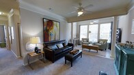 This single-floor residence at 427 offers a thoughtfully designed living space that balances comfort and style. The living area is a standout feature, showcasing elevated ceilings adorned with crown molding and decorative columns that lend an air of architectural refinement. A ceiling fan centered overhead provides both function and visual interest, while large windows along the far wall invite generous amounts of natural light, creating a warm and welcoming ambiance throughout the day. The living room is anchored by plush carpeting and offers ample space for a variety of furniture arrangements, as evidenced by the defined main seating zone and a separate sitting nook positioned near the windows — ideal for reading or casual conversation. The neutral color palette of the walls and flooring serves as a versatile backdrop, complemented by tasteful décor including bold artwork that adds a vibrant focal point. Architectural columns elegantly separate the main and secondary seating areas without fully closing off the open, airy feel of the space. Overall, this residence combines classic detailing with comfortable, livable design, making it a polished and inviting home.