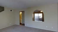 This two-floor space is designed with a clean, functional layout accented by thoughtful architectural details. The main living area is finished with wall-to-wall carpeting in a neutral tone, complementing the light beige walls that create a calm, understated ambiance throughout. A standout design feature is the arched pass-through or decorative niche set into one wall, framed with a contrasting darker surround and fitted with a ceiling light fixture. This arch frames a sliding glass door or window that opens to a view of an outdoor balcony or patio, allowing natural light to filter into the interior. Vertical blinds provide privacy and light control for this opening. Adjacent to the main living space, a hallway leads to a bathroom area, visible in the background, which features warm-toned cabinetry and a vanity with overhead lighting, suggesting a well-appointed and practical layout. The overall interior palette relies on neutral creams and beiges, contributing to a cohesive and welcoming atmosphere. The two-floor configuration provides additional living space and separation between areas, adding to the overall functionality of the property.