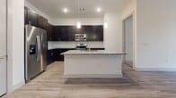 This thoughtfully designed single-floor residence showcases a contemporary aesthetic throughout, with consistent finishes that create a cohesive and polished living environment. The kitchen serves as a standout centerpiece, featuring rich dark espresso cabinetry that extends to the ceiling, a classic white subway tile backsplash, and striking granite countertops with a salt-and-pepper pattern. A large central island with an undermount sink and pendant lighting above provides both additional prep space and a natural gathering point. A full suite of stainless steel appliances, including a French door refrigerator, range, microwave, and dishwasher, completes the space. Light-toned wood-look flooring runs throughout the main living areas, lending warmth and continuity. The bedrooms are finished with plush carpeting and feature tall, narrow windows that bring in natural light. One bedroom includes sliding glass door access, adding an open, airy feel. The bathrooms are elegantly appointed with the same dark cabinetry and granite countertops found in the kitchen, large-format tile flooring, and tiled tub and shower surrounds. The primary bathroom offers a dual-sink vanity, a soaking tub, and direct access to a spacious walk-in closet with built-in shelving.