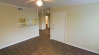 This two-floor space features a well-designed open layout that seamlessly connects the main living area to the kitchen. The living area is finished with rich, dark hardwood flooring that extends throughout, adding warmth and character to the space. Crown molding lines the ceiling, lending a polished, classic touch, while a ceiling fan with a pendant light provides both comfort and ambient lighting.

A pass-through counter connects the living area to the kitchen, allowing for easy interaction between spaces. The kitchen is visible through the opening, showcasing cabinetry with a warm wood finish and light-toned countertops, creating a functional and cohesive cooking area. Recessed lighting in the kitchen adds a clean, modern element.

The front entry features a solid white door that opens directly into the main living space, creating an immediate sense of openness upon arrival. Neutral-toned walls throughout provide a versatile backdrop, making the space easy to personalize. A hallway visible toward the rear of the unit leads further into the home, hinting at additional rooms across both floors. Overall, the space strikes a balance between classic architectural details and practical, everyday functionality.