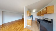 This single-floor residence showcases a clean, functional layout with warm parquet flooring running throughout the main living areas, lending a classic character to the space. The galley-style kitchen is efficiently designed, featuring honey-toned wood cabinetry that extends to the upper walls for generous storage, white countertops, and black appliances including a gas range and refrigerator. Tile flooring in the kitchen provides a practical contrast to the parquet found in the adjoining areas. A structural half-wall divides the kitchen from the living space, creating a sense of definition while maintaining an open feel.

The living area is notably bright, anchored by a large picture window that admits generous natural light and offers views of the surrounding neighborhood. The polished parquet flooring reflects this light, enhancing the airy quality of the room. A bedroom features the same parquet flooring, a sliding-door closet offering built-in storage, and direct access to additional rooms via a connecting hallway.

The bathroom presents a retro aesthetic with black-and-white mosaic tile flooring, square beige wall tiles accented with black border detail, a soaking tub with overhead shower, a pedestal-style sink, and brass hardware fixtures throughout.
