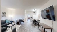 This single-floor unit offers a thoughtfully organized layout that maximizes every square foot across its four main areas: a combined kitchen and living room, a bedroom, a bathroom, and a hallway. The open-concept living and dining area is bright and airy, featuring light wood-look flooring, white walls, and ample room for both relaxing and studying. A wall-mounted television, a seating arrangement with dark upholstered sofas, accent wall art, and a dining table with industrial-style chairs give this shared space a modern, functional character. The kitchen is equipped with warm wood cabinetry, dark countertops, a stainless steel sink, and a full range, providing all the essentials for everyday cooking. The bedroom is generously sized and configured to accommodate multiple occupants, featuring loft-style bunk bed frames with integrated desks and storage drawers beneath — an efficient, space-conscious design well-suited for student living. The hallway provides a clean transition between rooms and includes a full-length mirror and louvered closet doors for additional storage. The bathroom features a full tub-shower combo with a crisp white surround, wood-look tile flooring, and simple, clean finishes throughout. The overall aesthetic is clean, minimal, and practical.