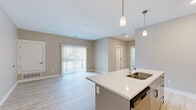 Located at 3800 E Campus Ave in Holland, Michigan, this single-floor unit offers three bedrooms and two bathrooms within a well-designed, efficient layout. The open-concept living and kitchen area serves as the heart of the home, featuring light wood-look luxury vinyl flooring, warm neutral-toned walls, and a sliding glass door that fills the living room with natural light and provides outdoor access. The kitchen is equipped with stainless steel appliances — including a refrigerator, range, and microwave — paired with crisp white cabinetry, a white quartz-topped island with a double-basin sink, and stylish pendant lighting overhead. The bedrooms are carpeted for comfort, with the primary bedroom offering a generous walk-in closet with wire shelving and a window overlooking greenery. The two bathrooms are finished with wood-look tile flooring; one features a full tub-shower combo while the other includes a stand-alone shower stall, each with a vanity and clean, neutral finishes. The cohesive design palette of soft beiges, whites, and light wood tones creates a bright, modern ambiance throughout the entire space.