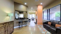 This single-story space is designed with an open-concept layout that promotes a seamless flow between living and dining areas. The polished concrete floors lend an industrial-chic aesthetic while remaining highly durable and easy to maintain. A prominent breakfast bar with stylish bar stools serves as a central gathering point, complemented by a pass-through window that connects to an adjacent kitchen area, adding both functionality and visual interest.

A distinctive orange accent column anchors the main living area, adding a bold pop of color against the neutral tan and beige wall tones. Recessed lighting throughout ensures even, well-distributed illumination across the space. Large windows along multiple walls flood the interior with natural light and offer views of a lush outdoor area, creating a bright and airy ambiance.

The dining area, visible in the background, is defined by a decorative area rug and features a table and chairs, making it ideal for gatherings. A leather sofa in the lounge area adds a comfortable and inviting touch. The overall design balances practicality with a warm, casual aesthetic, making this space well-suited for both everyday use and entertaining.