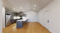 This single-story residence offers a thoughtfully designed layout accommodating four bedrooms, one full bathroom, a combined kitchen and living area, dedicated laundry, a closet, and a connecting hallway. The open-concept kitchen is a standout feature, showcasing gray shaker-style cabinetry, stainless steel appliances including a refrigerator, range, and microwave, white quartz countertops, a built-in sink island, and floating open shelves. Recessed lighting and vaulted ceilings enhance the sense of openness in the kitchen and living area, which is finished with warm-toned hardwood-style flooring. The living space includes a wall-mounted PTAC unit and direct access to an exterior door. The four bedrooms feature neutral carpet, ceiling fans, and windows that bring in natural light and wooded views. The full bathroom is cleanly appointed with gray cabinetry, a white countertop vanity, a glass-enclosed walk-in shower, and matching hardwood-style flooring. In-unit laundry is conveniently housed behind louvered doors, with a full-size washer and dryer included.