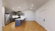 This single-story residence offers a thoughtfully designed layout accommodating four bedrooms, one full bathroom, a combined kitchen and living area, dedicated laundry, a closet, and a connecting hallway. The open-concept kitchen is a standout feature, showcasing gray shaker-style cabinetry, stainless steel appliances including a refrigerator, range, and microwave, white quartz countertops, a built-in sink island, and floating open shelves. Recessed lighting and vaulted ceilings enhance the sense of openness in the kitchen and living area, which is finished with warm-toned hardwood-style flooring. The living space includes a wall-mounted PTAC unit and direct access to an exterior door. The four bedrooms feature neutral carpet, ceiling fans, and windows that bring in natural light and wooded views. The full bathroom is cleanly appointed with gray cabinetry, a white countertop vanity, a glass-enclosed walk-in shower, and matching hardwood-style flooring. In-unit laundry is conveniently housed behind louvered doors, with a full-size washer and dryer included.