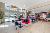 Located at 318 W Wilson Ave in Glendale, California, this single-floor community amenity space is a dynamic, artfully designed retreat that blends bold aesthetics with functional social areas. The main lounge is anchored by a full-size billiards table set atop a graphic patterned area rug, bathed in natural light from soaring floor-to-ceiling windows that overlook a resort-style outdoor pool. Oversized drum pendant lights hang above, lending a warm, inviting glow to the open-plan space. Built-in bookshelves line the walls, adding a curated, intellectual charm throughout.

A dedicated lounge area features a sleek wall-mounted fireplace framed by a striking white chevron-textured surround, complemented by plush navy accent chairs and a mounted flat-screen TV. Direct glass door access leads to a covered outdoor terrace with additional seating.

One of the standout features is a vibrant dining nook adorned with a bold, large-scale geometric mural in rich multicolor tones. A marble-topped round table surrounded by deep red chairs sits beneath a sculptural starburst chandelier, creating a truly artistic focal point. Nearby, a workspace counter finished in warm wood cabinetry is set against a refined herringbone-patterned wall. The space also connects to a colorful art installation corridor featuring a vivid mural and eclectic décor.
