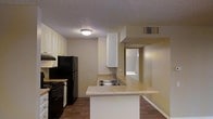 This single-story space centers around a practical, open-concept kitchen that serves as the heart of the layout. The kitchen is outfitted with white upper and lower cabinetry providing ample storage, black appliances including a refrigerator and gas range with an overhead exhaust hood, and a light-toned countertop peninsula that doubles as a workspace and casual dining area. The peninsula integrates a stainless steel double sink, enhancing both functionality and flow within the space.

Dark hardwood-style flooring runs throughout, offering a warm contrast to the light-colored walls and white cabinetry. A textured ceiling adds subtle visual detail, while a central flush-mount light fixture provides general illumination. An HVAC vent is visibly integrated into the ceiling, indicating a centralized climate control system.

The open layout allows sightlines into an adjoining room visible through a doorway at the rear, suggesting a connected, functional floor plan. The overall ambiance is clean and utilitarian, with a neutral color palette of warm beige tones creating a cohesive and understated interior atmosphere throughout the space.