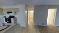 This single-floor space offers a practical and clean layout designed for comfortable everyday living. The open-concept design connects the kitchen and living areas seamlessly, creating a sense of spaciousness throughout. Light wood laminate flooring runs through the main living and hallway areas, adding warmth and continuity to the interior, while carpeting defines the living space.

The kitchen is positioned to the left and features light wood cabinetry with ample storage, a countertop workspace, and standard appliances including a stove. A textured ceiling runs throughout, giving the space a classic residential character.

A central hallway extends from the main living area toward the back of the unit, lined with a closet featuring sliding doors — ideal for additional storage. The hallway leads to a bright, naturally lit bathroom, visible at the end of the corridor, which benefits from a window that brings in natural light.

Neutral taupe and white wall tones create a calm, understated ambiance throughout the space. A floor vent provides in-floor heating, adding to the practical comfort of the unit. Overall, the space presents a tidy, functional interior with a warm and inviting atmosphere well-suited for a variety of uses.