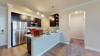 Located at 1703 Pope Rd in Bradenton, Florida, this single-story residence offers a well-designed layout with a cohesive blend of contemporary finishes and practical living spaces. The open-concept kitchen serves as the focal point of the home, featuring rich dark espresso cabinetry, stainless steel appliances including a refrigerator and microwave, light quartz countertops, and a peninsula with pendant lighting overhead. The kitchen transitions seamlessly into the living area, which is generously sized, carpeted, and outfitted with crown molding and a ceiling fan, creating a comfortable and airy atmosphere. Natural light filters in through multiple windows dressed with blinds, brightening the neutral-toned walls throughout. The bedrooms are spacious and carpeted, each offering closet storage and windows that allow in natural light. The bathroom is well-appointed with dark vanity cabinetry, a white countertop, a large mirror, and a tub-shower combination with wood-look flooring. A dedicated laundry area houses a full-size Samsung washer and dryer set with overhead shelving for added storage. Wood-look plank flooring runs through the main living and kitchen areas, complementing the overall clean, modern aesthetic of the home.