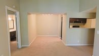 This single-floor residence offers a clean, open layout designed for comfortable everyday living. The main living area is finished with soft, neutral-toned wall-to-wall carpeting that adds warmth underfoot, while crisp white baseboards frame the space with a polished touch. The walls are painted in a light, muted green tone that lends a calm and airy ambiance throughout.

A defined recessed alcove within the living area is illuminated by a row of recessed ceiling lights, creating a naturally framed zone ideal for a dining or secondary seating arrangement. The open passage connecting this alcove to the adjacent areas allows for an easy, flowing floor plan.

To one side, a kitchen is partially visible, showcasing dark wood cabinetry paired with a light-toned countertop that extends to form a pass-through bar — a practical and social feature that connects the kitchen to the living space. A black appliance is visible within the kitchen, adding contrast to the overall neutral palette.

The space benefits from a spacious, uncluttered feel, with high walls that enhance the sense of volume throughout. The single-floor layout ensures convenient accessibility, and the cohesive neutral color scheme throughout creates a versatile backdrop well-suited to a variety of interior styles.