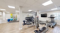 Located in Berry Hill, TN, this dedicated fitness facility offers a thoughtfully designed exercise space that accommodates a full range of workout activities. The open floor plan is finished with light wood-look flooring throughout, lending a clean and welcoming aesthetic that complements the bright, neutral-toned walls and white trim.

The space is illuminated by recessed panel lighting set within a drop-tile ceiling, ensuring consistent, even brightness across the entire floor. Structural columns are neatly integrated into the layout, subtly defining different workout zones without disrupting the open feel.

The strength training area is well-appointed with a multi-station cable machine, a tiered dumbbell rack with a full range of free weights, a flat workout bench, and additional resistance equipment. A wall-mounted flat-screen TV overlooks this zone, providing entertainment during workouts. The cardio section features an upright stationary bike and a rowing machine, while a dedicated open floor area with foam floor mats offers space for stretching, yoga, or functional training. Stability balls and resistance accessories are neatly stored along the wall.

A utility area with white cabinetry, granite-style countertops, and a sink provides practical support for the space. Multiple access doors offer convenient entry and exit points throughout the facility.
