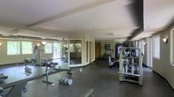 This fitness center offers a comprehensive workout environment designed to accommodate a variety of exercise routines. The space is generously sized, with a open floor plan that allows for easy movement between equipment stations. Large mirrors line one wall, visually expanding the room and allowing users to monitor their form during workouts. Natural light filters in through windows that overlook a green outdoor area, creating a bright and energizing atmosphere. The flooring features durable rubber matting, ideal for both cardio and strength training activities. A selection of cardio machines, including ellipticals and treadmills, are arranged along one side of the room, while free weights and a weight bench are conveniently positioned nearby. Additional strength-training equipment and a flat-screen display round out the facility's offerings. The textured ceiling and neutral-toned walls contribute to a clean, functional aesthetic throughout. A ceiling fan provides added ventilation and comfort during intense workouts. Access doors are visible along the back wall, providing convenient entry and exit points. Overall, this fitness center is thoughtfully laid out to support a well-rounded exercise regimen in a comfortable, well-lit setting.
