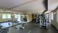 This fitness center offers a comprehensive workout environment designed to accommodate a variety of exercise routines. The space is generously sized, with a open floor plan that allows for easy movement between equipment stations. Large mirrors line one wall, visually expanding the room and allowing users to monitor their form during workouts. Natural light filters in through windows that overlook a green outdoor area, creating a bright and energizing atmosphere. The flooring features durable rubber matting, ideal for both cardio and strength training activities. A selection of cardio machines, including ellipticals and treadmills, are arranged along one side of the room, while free weights and a weight bench are conveniently positioned nearby. Additional strength-training equipment and a flat-screen display round out the facility's offerings. The textured ceiling and neutral-toned walls contribute to a clean, functional aesthetic throughout. A ceiling fan provides added ventilation and comfort during intense workouts. Access doors are visible along the back wall, providing convenient entry and exit points. Overall, this fitness center is thoughtfully laid out to support a well-rounded exercise regimen in a comfortable, well-lit setting.