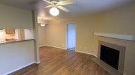This single-floor residence offers a welcoming and functional layout designed for comfortable everyday living. The main living area is finished with rich, warm-toned hardwood-style flooring that extends throughout, adding a sense of warmth and continuity to the space. A ceiling fan with a built-in light fixture is centrally positioned, providing both air circulation and ambient lighting. A decorative pendant light hangs nearby, adding a subtle accent to the open area. One of the standout features of the living room is the fireplace, complete with a clean, simple mantle, serving as a natural focal point for the room. Soft yellow-toned walls contribute to the bright and airy feel of the interior. A partial wall or breakfast bar divides the living area from an adjacent space, hinting at an open-concept connection to the kitchen or dining area. A doorway leads to additional rooms, suggesting a practical and well-organized floor plan. White trim and baseboards provide a crisp contrast against the warm wall tones, giving the space a polished, finished appearance throughout. Overall, the residence presents a cozy yet spacious atmosphere well-suited for a variety of living arrangements.