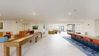 This expansive single-floor amenity lounge offers a thoughtfully designed communal space that blends recreation, relaxation, and social interaction. The space is anchored by a dramatic double-height entrance lobby, where soaring ceilings and a striking floor-to-ceiling decorative feature wall create a bold architectural statement. Large windows and glass doors throughout flood the interior with abundant natural light and provide views of the surrounding exterior.

The open floor plan flows seamlessly across light-toned terrazzo tile floors, transitioning to plush blue area rugs that define distinct activity zones. A dedicated game room houses a full-size billiard table and a regulation shuffleboard table, both illuminated by pendant lighting. A long bar-height wooden counter with vibrant red stools serves as a social hub, complemented by a wall-mounted television and a tiled wood-look accent wall nearby.

Comfortable lounge seating areas are furnished with contemporary chairs, sofas, and accent tables, accented by colorful artwork on white walls. Built-in banquette seating adds intimate gathering spots throughout. The space also features a café-style counter area near the entrance, ideal for casual work or conversation. Recessed lighting throughout ensures a clean, modern aesthetic across every zone of this versatile communal space.