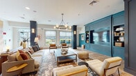 This single-floor community space in Kingston, Massachusetts, presents a carefully curated layout spanning several interconnected areas, each defined by its own character while maintaining a cohesive design language throughout. Light hardwood flooring runs continuously across the space, grounding each zone in warmth and continuity.

The main lounge area serves as a central gathering point, anchored by a bold teal built-in entertainment wall with integrated shelving, flanked by dark navy columns. A contemporary multi-globe chandelier illuminates an arrangement of plush sofas, leather accent chairs, and layered area rugs. Large windows invite natural light and outdoor views, enhancing the airy atmosphere.

Adjacent transitional seating areas feature white upholstered chairs, barn-style sliding glass doors, and curated artwork, creating quieter spaces for conversation or relaxation. A dining nook includes a round wood table beneath a pendant light, set against textured grasscloth wallcovering and a built-in display cabinet in a soft sage finish.

The kitchen area is appointed with natural wood cabinetry, stone countertops, vertical shiplap walls in muted sage, open floating shelves, and a central island with bar seating beneath globe pendant lights. The game room offers a distinctly different ambiance, featuring an ornate coffered ceiling, billiards table, foosball, shuffleboard, leather booth seating, and wall-mounted televisions.