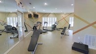 This single-floor fitness center offers a clean, functional space designed to support a variety of workout routines. The room is decorated with a distinctive geometric accent pattern in warm orange and tan tones across the walls, adding visual energy to the exercise environment. Recessed lighting illuminates the space from above, complemented by natural light streaming through multiple white-shuttered windows that line the walls. Large wall-mounted mirrors along one side of the room expand the sense of space and allow exercisers to monitor their form during workouts. The flooring features durable, cushioned material well-suited for fitness activities. Cardio equipment, including treadmills and an elliptical trainer, is arranged to maximize floor space and airflow. Flat-screen televisions are mounted on the wall, providing entertainment during workouts. Adjustable weight benches are also available for strength training. The overall ambiance is bright, motivating, and practical, with a layout that balances equipment accessibility and open movement space, making it a convenient and inviting facility for daily exercise.