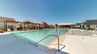 This expansive two-floor resort-style amenity complex centers around a generously sized swimming pool with crystal-clear turquoise water, designed to serve as a community gathering destination. The pool features a shallow sun shelf area equipped with in-water lounge chairs and colorful accent cushions, ideal for relaxing at the water's edge. Surrounding the pool is a broad concrete and stone deck furnished with multiple chaise loungers and navy blue market umbrellas, providing ample space for sunbathing under open skies.

The complex includes several architectural structures that frame the pool area. A modern clubhouse building features warm wood-panel exterior cladding, a standing-seam metal roof, and large glass doors that open directly to the pool deck. String lights overhead add a welcoming ambiance. Multiple pergola structures with slatted wood roofing and metal frames offer shaded seating areas with patio furniture, creating comfortable outdoor gathering spots. The surrounding community features cottage-style residential buildings with gabled roofs in a variety of exterior colors, lending a cohesive neighborhood aesthetic. Mature landscaping, including young trees and manicured greenery, softens the hardscape throughout the space.