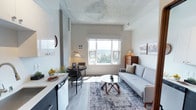 Located at 229 Andover Park E in Seattle, WA, this single-floor unit offers a thoughtfully designed open layout that maximizes both function and style. The living area is anchored by light hardwood-style flooring and a distinctive exposed concrete ceiling that adds an industrial character to the space. A large window floods the room with natural light and frames sweeping views of the surrounding treetops and cityscape, creating a bright and airy ambiance throughout.

The kitchen is outfitted with a full suite of stainless steel appliances, including a refrigerator, electric range, and over-the-range microwave. Sleek two-tone cabinetry — crisp white uppers paired with dark lower cabinets — complements the clean white countertops and undermount stainless steel sink, delivering a modern and efficient cooking space.

The living area comfortably accommodates both a seating and dining arrangement, with the open floor plan creating a seamless flow between the two. A sliding-door closet provides practical storage within the main living space. The bathroom is clean and simply appointed, featuring tile flooring and tasteful framed wall art. Overall, this unit combines contemporary finishes with functional design in a compact, well-appointed package.