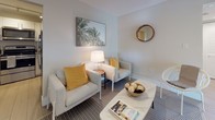 This well-appointed single-floor residence showcases a clean, contemporary design with a cohesive neutral palette and light wood-style flooring that flows seamlessly throughout the living areas and bedrooms. The modern kitchen is outfitted with white cabinetry, grey quartz countertops, a grey tile backsplash, and a full suite of stainless steel appliances, including a range, over-the-range microwave, and dishwasher — all arranged in an efficient U-shaped layout. The living area offers an intimate and stylish setting, featuring a patterned area rug, a marble-top coffee table, and warm accent tones that complement the soft grey walls. Multiple bedrooms are present, each designed with simplicity and functionality in mind. One bedroom features floor-to-ceiling sliding glass doors that open directly to a private outdoor terrace with lush tropical views, accompanied by a ceiling fan and generous closet space, including a dedicated walk-in closet with wire shelving. Additional bedrooms are served by natural light through roller-shade windows and include built-in closet storage. The bathroom is finished with large-format white tile, a glass-enclosed soaking tub and shower combination, and a wood-toned vanity. Light, airy hallways connect each space, contributing to an open and easy-flowing floor plan throughout.
