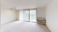 This single-floor unit offers a straightforward, functional layout across several distinct areas. The living area is generously sized, featuring light beige wall-to-wall carpeting, neutral white walls, and a textured tile ceiling. A large sliding glass door fitted with vertical blinds serves as the focal point of the living space, allowing abundant natural light and offering outdoor views of surrounding greenery. A wall-mounted HVAC unit and track lighting add practical utility to the room.

The bedrooms continue the same carpeted flooring and neutral palette. Each room benefits from natural light via windows dressed with vertical blinds. One bedroom features dual bifold closet doors providing ample built-in storage with shelving. The rooms connect to a hallway with additional storage access.

The unit includes two bathrooms. One features white square tile wainscoting, a vanity with mirror, and a combined tub-shower. The second bathroom similarly incorporates white tile wainscoting, a vanity with a stone-patterned countertop, a toilet, and a separate tub, with large-format tile flooring throughout.

The kitchen, visible in the floor plan, is positioned at one end of the unit and includes cabinetry and appliances. Overall, the space presents a clean, neutral, and move-in-ready environment.