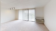 This single-floor unit offers a straightforward, functional layout across several distinct areas. The living area is generously sized, featuring light beige wall-to-wall carpeting, neutral white walls, and a textured tile ceiling. A large sliding glass door fitted with vertical blinds serves as the focal point of the living space, allowing abundant natural light and offering outdoor views of surrounding greenery. A wall-mounted HVAC unit and track lighting add practical utility to the room.

The bedrooms continue the same carpeted flooring and neutral palette. Each room benefits from natural light via windows dressed with vertical blinds. One bedroom features dual bifold closet doors providing ample built-in storage with shelving. The rooms connect to a hallway with additional storage access.

The unit includes two bathrooms. One features white square tile wainscoting, a vanity with mirror, and a combined tub-shower. The second bathroom similarly incorporates white tile wainscoting, a vanity with a stone-patterned countertop, a toilet, and a separate tub, with large-format tile flooring throughout.

The kitchen, visible in the floor plan, is positioned at one end of the unit and includes cabinetry and appliances. Overall, the space presents a clean, neutral, and move-in-ready environment.