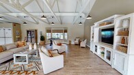 This single-story space is defined by its striking architectural centerpiece: soaring vaulted ceilings with fully exposed, white-painted wood trusses that span the entire open floor plan. Black industrial-style pendant lights hang from the trusses, complementing the warm, light-toned hardwood-style flooring that runs throughout. The expansive main gathering area is furnished with neutral linen seating arrangements, a patterned area rug, and a large built-in white entertainment center housing a flat-screen television. A foosball table and wicker accent chairs provide additional recreational options, while multiple windows fitted with woven shades allow generous natural light to fill the room. Adjacent to the main area, a well-appointed kitchen features crisp white shaker cabinetry, a long stainless steel sink set into a generous island, open rustic wood shelving accented by decorative patterned tile, and the same vaulted truss ceiling continuing overhead. A separate office or leasing nook offers a long wooden work desk, a decorative mirror board displaying floor plans, and vibrant coral accent chairs beside a window, creating a welcoming and functional workspace. The overall ambiance blends farmhouse warmth with an open, airy character throughout.