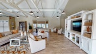 This single-story space is defined by its striking architectural centerpiece: soaring vaulted ceilings with fully exposed, white-painted wood trusses that span the entire open floor plan. Black industrial-style pendant lights hang from the trusses, complementing the warm, light-toned hardwood-style flooring that runs throughout. The expansive main gathering area is furnished with neutral linen seating arrangements, a patterned area rug, and a large built-in white entertainment center housing a flat-screen television. A foosball table and wicker accent chairs provide additional recreational options, while multiple windows fitted with woven shades allow generous natural light to fill the room. Adjacent to the main area, a well-appointed kitchen features crisp white shaker cabinetry, a long stainless steel sink set into a generous island, open rustic wood shelving accented by decorative patterned tile, and the same vaulted truss ceiling continuing overhead. A separate office or leasing nook offers a long wooden work desk, a decorative mirror board displaying floor plans, and vibrant coral accent chairs beside a window, creating a welcoming and functional workspace. The overall ambiance blends farmhouse warmth with an open, airy character throughout.