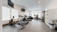 Located at 5707 S Spotted Rd in Spokane, WA, this dedicated fitness room offers a well-organized and fully equipped workout environment within a compact yet efficiently utilized single-level footprint. The space features a clean, modern design with crisp white walls, recessed lighting, and durable speckled rubber flooring throughout, providing both functionality and a polished aesthetic suited for regular exercise use.

Natural light enters through a row of transom-style windows positioned high along one wall, brightening the interior without compromising usable wall and floor space. A wall-mounted flat-screen television is positioned for visibility across the main workout area, with an additional ceiling-mounted monitor near the entry side of the room.

The cardio zone is anchored by True-brand treadmills, an elliptical trainer, and a stair climber, arranged to allow comfortable circulation between machines. The strength training area includes a multi-functional cable machine with an adjustable bench, a rack of dumbbells in graduated weights, a weight bench with medicine balls, and a cable/weight stack system. Built-in open shelving units along one wall provide organized storage for accessories and equipment. A water fountain and entry door complete the space, supporting practical day-to-day use of the facility.