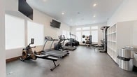 Located at 5707 S Spotted Rd in Spokane, WA, this dedicated fitness room offers a well-organized and fully equipped workout environment within a compact yet efficiently utilized single-level footprint. The space features a clean, modern design with crisp white walls, recessed lighting, and durable speckled rubber flooring throughout, providing both functionality and a polished aesthetic suited for regular exercise use.

Natural light enters through a row of transom-style windows positioned high along one wall, brightening the interior without compromising usable wall and floor space. A wall-mounted flat-screen television is positioned for visibility across the main workout area, with an additional ceiling-mounted monitor near the entry side of the room.

The cardio zone is anchored by True-brand treadmills, an elliptical trainer, and a stair climber, arranged to allow comfortable circulation between machines. The strength training area includes a multi-functional cable machine with an adjustable bench, a rack of dumbbells in graduated weights, a weight bench with medicine balls, and a cable/weight stack system. Built-in open shelving units along one wall provide organized storage for accessories and equipment. A water fountain and entry door complete the space, supporting practical day-to-day use of the facility.