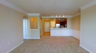 This single-floor space presents an open-concept design that seamlessly connects the living area to the kitchen. The living area is finished with plush, neutral-toned carpeting and features a decorative arched wall niche, adding a touch of architectural character to the space. Crown molding runs along the ceiling, lending a polished, refined feel throughout.

The kitchen is positioned at the back of the open layout and is defined by a white wainscoted breakfast bar that serves as a natural divider between the kitchen and living areas. Rich, warm-toned cabinetry lines the kitchen walls, complemented by a stainless steel refrigerator and a stainless steel sink. Track lighting illuminates both the kitchen and the transition area, ensuring ample light across the space.

A hallway visible from the main living area leads to additional rooms, with an arched doorway adding continuity to the architectural detailing found throughout. A secondary door at the end of the hallway provides access to what appears to be a utility or storage space. The overall ambiance is warm, clean, and functional, with consistent neutral tones and classic architectural accents throughout.