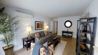 This single-floor space centers around a thoughtfully designed living area that combines comfort and style through a cohesive neutral color palette of grays, whites, and warm wood tones. The living room is furnished with a gray upholstered sofa and coordinating accent chairs, both adorned with bold orange-and-white patterned cushions that add visual interest to the space. A rectangular coffee table anchors the seating arrangement atop a plush area rug, while hardwood-style flooring extends throughout the room, lending warmth and continuity.

The walls are painted in a clean, light gray tone, complemented by white crown molding and baseboards. A framed piece of abstract artwork hangs prominently on one wall, while a circular black-framed mirror serves as a decorative focal point on the opposite wall above a sleek console table. A wall-mounted air conditioning unit is visible, providing climate control. Table lamps with warm-toned shades offer ambient lighting throughout the space.

A doorway toward the rear of the living area leads to additional rooms, suggesting a practical and connected layout. Overall, the space presents a modern, well-organized interior with a welcoming ambiance.