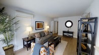 This single-floor space centers around a thoughtfully designed living area that combines comfort and style through a cohesive neutral color palette of grays, whites, and warm wood tones. The living room is furnished with a gray upholstered sofa and coordinating accent chairs, both adorned with bold orange-and-white patterned cushions that add visual interest to the space. A rectangular coffee table anchors the seating arrangement atop a plush area rug, while hardwood-style flooring extends throughout the room, lending warmth and continuity.

The walls are painted in a clean, light gray tone, complemented by white crown molding and baseboards. A framed piece of abstract artwork hangs prominently on one wall, while a circular black-framed mirror serves as a decorative focal point on the opposite wall above a sleek console table. A wall-mounted air conditioning unit is visible, providing climate control. Table lamps with warm-toned shades offer ambient lighting throughout the space.

A doorway toward the rear of the living area leads to additional rooms, suggesting a practical and connected layout. Overall, the space presents a modern, well-organized interior with a welcoming ambiance.