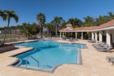 Located at Greens Edge in Fort Myers, Florida, this two-story community pool facility offers a resort-style outdoor retreat designed for relaxation and recreation. The centerpiece is a generously sized swimming pool with crystal-clear turquoise water, framed by a broad travertine deck that provides ample space for lounging. Surrounding the pool, rows of chaise lounge chairs and seating areas invite residents to soak up the Florida sunshine.

The pool building itself showcases Mediterranean-inspired architecture, featuring a distinctive terracotta tile roof, stucco exterior, and classic columned covered walkways that offer shaded gathering areas along the pool's edge. French doors provide access to interior amenities, while the covered pavilion areas accommodate outdoor dining furniture and additional seating.

Lush tropical landscaping, including towering palm trees, frames the entire facility, enhancing the resort ambiance. A dedicated spa or hot tub is positioned adjacent to the main pool, offering a relaxing complement to the swim area. The facility also features a decorative hedge wall with a "Good Vibes Only – Greens Edge" sign, adding a welcoming, community-focused touch. Views of the surrounding residential buildings and a scenic lake backdrop complete this vibrant outdoor amenity space.