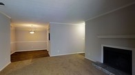 This single-floor home offers a clean, classic interior defined by thoughtful architectural details and a neutral color palette throughout. The living room serves as a central gathering space, featuring wall-to-wall carpeting, a traditional fireplace with a white mantel surround, and soft gray walls that create a calm, inviting atmosphere. Crown molding trims the ceiling, adding a refined touch to the overall design. Adjacent to the living room, the dining area transitions to rich hardwood flooring, providing a warm contrast to the carpeted living space. A pendant light fixture anchors the dining area, offering both functional illumination and a subtle decorative accent. The open sightline between the two spaces enhances the sense of flow and connectivity, making the layout feel spacious and well-connected. White baseboards and trim work are consistent throughout, contributing to a cohesive and polished look. The muted tones of the walls and flooring create a versatile backdrop well-suited to a variety of furnishing styles. Overall, the space combines traditional architectural character with a straightforward, functional layout ideal for comfortable everyday living.
