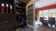 This professional office space is thoughtfully designed to support both focused individual work and collaborative meetings. The layout is divided into two distinct functional areas connected by a frosted-glass panel door, creating a sense of privacy while maintaining an open flow between spaces.

The private workstation area is anchored by a floor-to-ceiling dark espresso wood built-in unit, complete with integrated shelving, a built-in desk, and cabinetry with brass hardware. Decorative accents including red ceramic vases, candle holders, and curated books lend a refined, executive character to the space. Patterned carpet flooring adds texture underfoot, and warm taupe walls provide a neutral backdrop that highlights the richness of the woodwork.

The adjacent conference room makes a bold visual statement with deep crimson accent walls and floor-length neutral drapes framing a large multi-pane window. The window fills the room with natural light and offers views of mature trees and landscaping beyond. A substantial oval conference table surrounded by sleek, modern black office chairs serves as the centerpiece, making the room well-suited for professional meetings and presentations.

Throughout both areas, the design balances classic architectural detailing — including crown molding and paneled millwork — with contemporary furnishings, creating a polished and functional workspace.