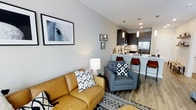 This single-floor space showcases a thoughtfully designed open-concept layout that seamlessly connects the living area to the kitchen, creating a cohesive and functional environment. The living area features warm wood-tone flooring and is furnished with a mustard leather sofa and a gray accent chair, accented by bold geometric-patterned throw pillows and a textured area rug. Curated wall art adds a contemporary artistic touch to the light gray walls, enhancing the modern aesthetic. Recessed lighting throughout the space provides a clean, well-lit atmosphere. The kitchen is finished with dark espresso cabinetry, pendant lighting, and stainless steel appliances, including a full-size refrigerator. A white countertop island with bar seating — fitted with sleek red and natural-toned bar stools — serves as both a functional workspace and a casual dining spot. The overall ambiance is modern and inviting, with a neutral color palette punctuated by warm accent tones. The open flow between living and culinary spaces makes this unit well-suited for both everyday living and entertaining.