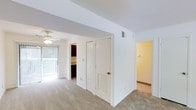 This two-floor residence offers a clean, functional layout designed for comfortable everyday living. The interior is finished in crisp white throughout, creating a bright and airy atmosphere complemented by neutral-toned carpeting that extends across the main living areas.

A central hallway connects the primary living spaces, with white panel doors providing access to storage closets, including a louvered-door closet ideal for utility or linen storage. The living area benefits from an abundance of natural light streaming through a sliding glass door that leads to an outdoor space, framed by vertical blinds. A ceiling fan with a light fixture is positioned overhead, ensuring both air circulation and ambient lighting.

Adjacent to the main living area, a partially visible room features dark wood cabinetry, suggesting a dedicated workspace or built-in storage nook. A warm yellow-toned hallway leads further into the unit, connecting to additional rooms across both floors.

The overall design emphasizes practicality and openness, with a neutral color palette, adequate storage solutions, and a layout that facilitates easy movement between living spaces. HVAC vents are integrated into the ceiling, ensuring year-round climate comfort throughout the home.