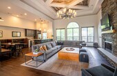 This well-appointed single-floor space blends rustic warmth with contemporary sophistication throughout its open, flowing layout. The main lounge commands immediate attention with soaring ceilings adorned by coffered panels clad in reclaimed wood, anchored by a striking multi-arm chandelier that casts a warm glow over the generous seating area below. Large arched and rectangular windows flood the space with natural light, framing views of the surrounding greenery. A floor-to-ceiling stacked-stone fireplace serves as a dramatic focal point, paired with a wood mantel, wall-mounted television, and built-in wooden shelving curated with books and décor. Rich hardwood flooring runs throughout, grounding the space with earthy warmth. Adjacent to the lounge, a dining nook features leather booth seating and distinctive geometric wood wall art, creating an intimate setting for gathering. The kitchen is a standout, outfitted with deep navy shaker-style cabinetry, gold hardware, a subway tile backsplash, stainless steel appliances, and a sleek quartz peninsula with bar stool seating. Pendant lighting and an open layout connect the kitchen seamlessly to the surrounding social areas, making this space ideal for community engagement and everyday comfort.