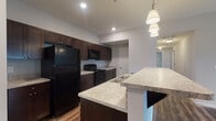 Located at 5695 Cherry Bottom Rd in Columbus, Ohio, this single-floor residence offers a well-designed open-concept layout with consistent, modern finishes throughout. The kitchen serves as a central highlight, featuring rich dark espresso cabinetry, light granite countertops, and a full suite of black appliances including a refrigerator, range, and over-the-range microwave. A peninsula with an extended granite overhang provides additional prep space and casual seating, while pendant lighting and recessed ceiling lights illuminate the area effectively. Durable wood-style flooring runs throughout the main living areas and kitchen, adding warmth and cohesion to the space. The living area is spacious and open, with a ceiling fan and access to natural light. The bedrooms are carpeted for comfort, with clean white walls and convenient access to the bathrooms. The bathrooms mirror the kitchen's design language, featuring matching dark vanity cabinetry, granite-topped sinks, and gray-toned walls. One bathroom includes a walk-in glass-door shower, while the other is equipped with a shower-tub combination. Overall, the unit presents a cohesive, contemporary aesthetic with practical finishes suited for everyday living.