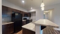 Located at 5695 Cherry Bottom Rd in Columbus, Ohio, this single-floor residence offers a well-designed open-concept layout with consistent, modern finishes throughout. The kitchen serves as a central highlight, featuring rich dark espresso cabinetry, light granite countertops, and a full suite of black appliances including a refrigerator, range, and over-the-range microwave. A peninsula with an extended granite overhang provides additional prep space and casual seating, while pendant lighting and recessed ceiling lights illuminate the area effectively. Durable wood-style flooring runs throughout the main living areas and kitchen, adding warmth and cohesion to the space. The living area is spacious and open, with a ceiling fan and access to natural light. The bedrooms are carpeted for comfort, with clean white walls and convenient access to the bathrooms. The bathrooms mirror the kitchen's design language, featuring matching dark vanity cabinetry, granite-topped sinks, and gray-toned walls. One bathroom includes a walk-in glass-door shower, while the other is equipped with a shower-tub combination. Overall, the unit presents a cohesive, contemporary aesthetic with practical finishes suited for everyday living.